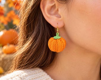May include: A crochet pumpkin earring featuring a bright orange spherical body with vertical ribbed texture and a small green stem at the top. The pumpkin is attached to a gold-toned metal hook finding that hangs from the earlobe.