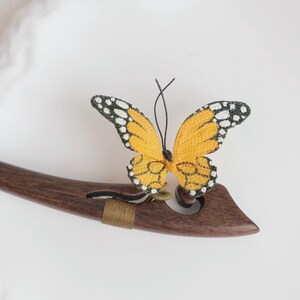 May include: A decorative butterfly hair accessory. The butterfly has yellow wings with black and white detailing, and is attached to a dark brown wooden hair stick. The hair stick has a curved design.