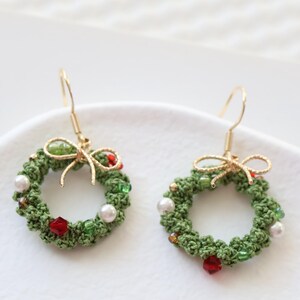 May include: A pair of green crocheted Christmas wreath earrings with red and white beads, gold bows, and gold hooks.