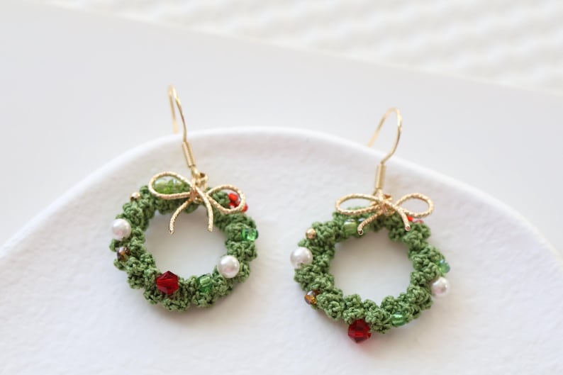 May include: A pair of green crocheted Christmas wreath earrings with red and white beads, gold bows, and gold hooks.