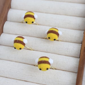 May include: Four handmade rings with crochet bee designs. Each bee has a yellow body with brown stripes, white wings, and a gold-colored ring band. The rings are displayed on a cream-colored fabric.
