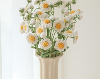 May include: A bouquet of crocheted white daisies with yellow centers in a cream-colored vase. The vase is on a white table with a book and a card that says "Little Crochet Flower by Layla" and "Wishes for you".