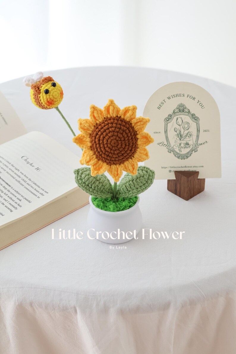 May include: A handmade crochet sunflower with a brown center and yellow petals, in a white pot with green leaves. A crochet bee is on a stem. A card with text "Best Wishes For You" is in the background. The text "Little Crochet Flower" is at the bottom.