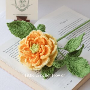 K&ouml;nnte beinhalten: Eine handgefertigte H&auml;kelblume mit gelben und orangenen Bl&uuml;tenbl&auml;ttern und gr&uuml;nen Bl&auml;ttern. Die Blume liegt auf einem offenen Buch. Der Stiel und die Bl&auml;tter sind gr&uuml;n, und die Blume hat ein detailliertes, strukturiertes Aussehen. Der Text "Little Crochet Flower" ist sichtbar.
