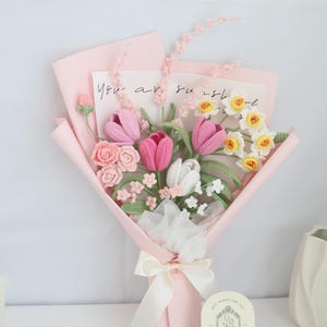 May include: A pink and white crocheted flower bouquet with a card that says "You are sunshine". The bouquet is wrapped in pink tissue paper and tied with a white ribbon.