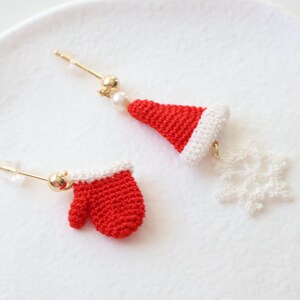 May include: A pair of crocheted Christmas earrings. One earring features a red mitten and the other features a white snowflake and a red and white Santa hat.