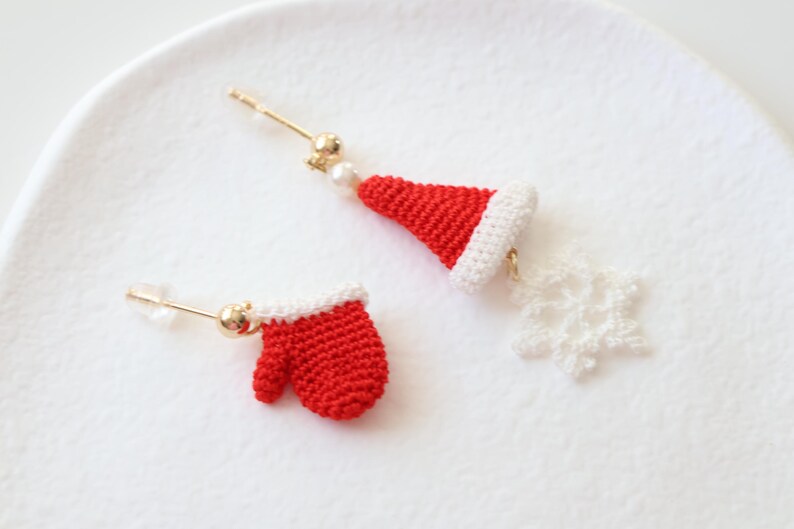 May include: A pair of crocheted Christmas earrings. One earring features a red mitten and the other features a white snowflake and a red and white Santa hat.