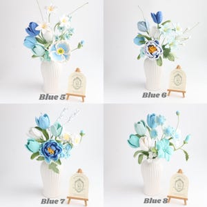 May include: Four arrangements of crocheted flowers in white ribbed vases, labeled Blue 5, Blue 6, Blue 7, and Blue 8. Each bouquet features various shades of blue, white, and green yarn petals, leaves, and stems, displayed with small wooden easels.