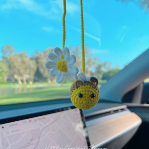 May include: A crocheted bee and daisy hanging from a car's rearview mirror. The bee is yellow with black stripes and brown antennae. The daisy is white with a yellow center. The background is a blue sky and green trees.