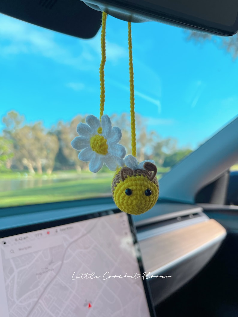 May include: A crocheted bee and daisy hanging from a car's rearview mirror. The bee is yellow with black stripes and brown antennae. The daisy is white with a yellow center. The background is a blue sky and green trees.