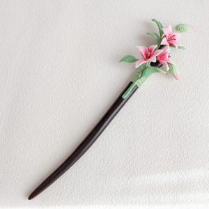 May include: A dark brown wooden hair stick with a decorative floral design. The flowers are pink with green leaves and accents. The hair accessory is a unique and elegant addition to any hairstyle.