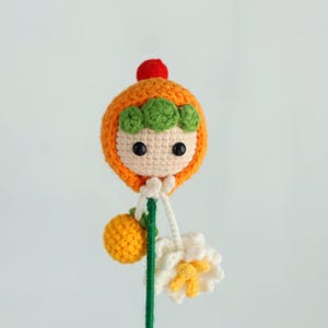 Puede incluir: Mu&ntilde;eca de ganchillo hecha a mano con capucha naranja, detalles verdes y un pomp&oacute;n rojo. La mu&ntilde;eca tiene una cara blanca con ojos negros. Sostiene una fruta amarilla de ganchillo y una flor blanca con detalles amarillos.