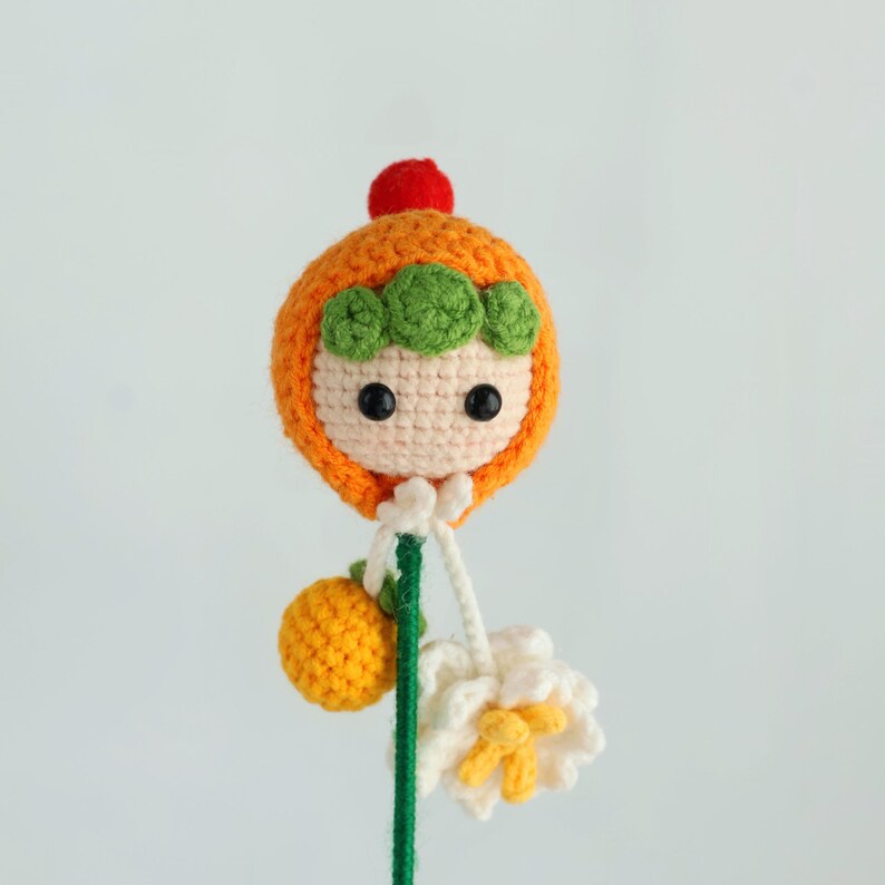 Puede incluir: Mu&ntilde;eca de ganchillo hecha a mano con capucha naranja, detalles verdes y un pomp&oacute;n rojo. La mu&ntilde;eca tiene una cara blanca con ojos negros. Sostiene una fruta amarilla de ganchillo y una flor blanca con detalles amarillos.