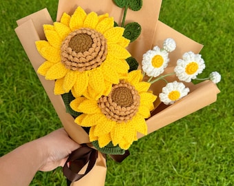 May include: A handmade bouquet of crocheted flowers, featuring two large yellow sunflowers with brown centers, white daisies, and green leaves. The flowers are wrapped in brown paper and tied with a brown ribbon. The bouquet is held against a background of green grass.