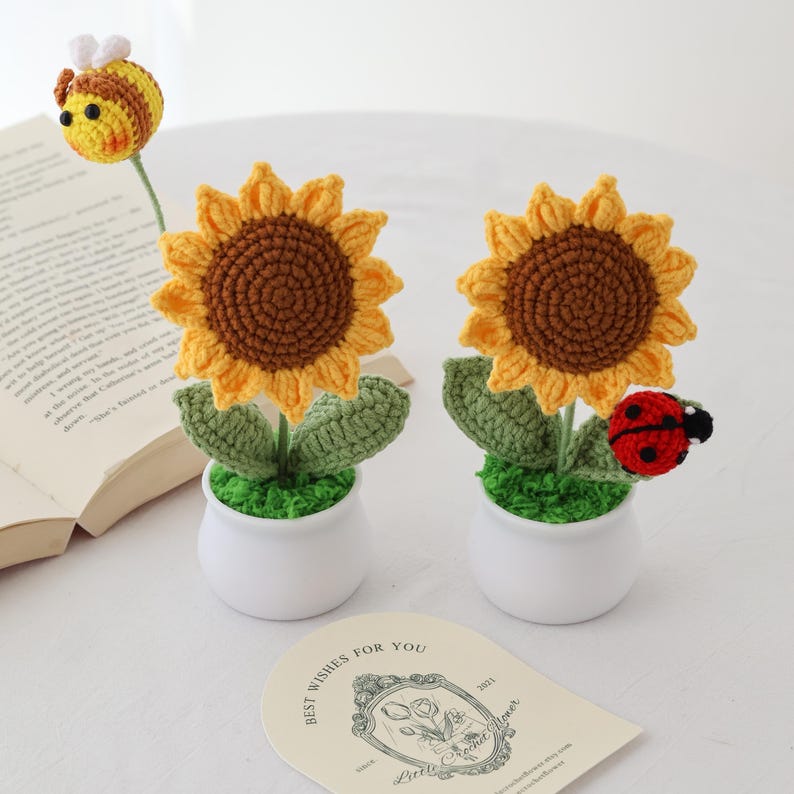 May include: Two crochet sunflowers with yellow petals and brown centers sit in white pots with green 'grass'. A crochet ladybug and bee are also present. A card with the text "BEST WISHES FOR YOU" is in the foreground. The sunflowers are handmade.