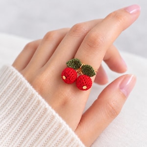May include: A ring featuring two small, round, red crocheted spheres resembling cherries, each topped with a tiny gold-toned accent. Two dark green crocheted leaves are attached to the top of the cherries, which are mounted on a thin gold-toned metal band.