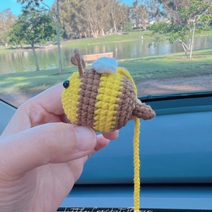 May include: A crocheted bee toy with brown and yellow stripes, a white crocheted head, and a long yellow yarn tail. The bee is being held by a hand in front of a blurry background of a lake and trees.
