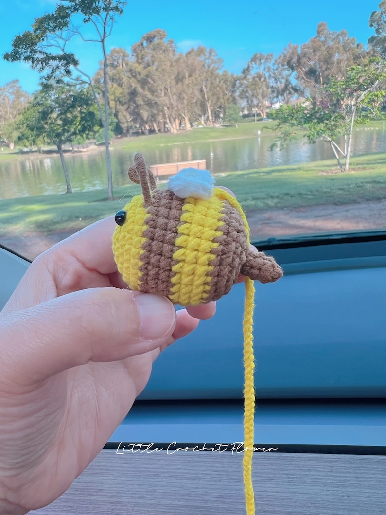 May include: A crocheted bee toy with brown and yellow stripes, a white crocheted head, and a long yellow yarn tail. The bee is being held by a hand in front of a blurry background of a lake and trees.