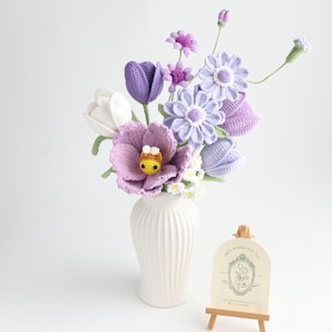 May include: A bouquet of crocheted flowers in shades of white, lavender, and purple, featuring tulips, daisies, and a small yellow and brown bee in the center. The flowers are arranged in a white ribbed vase next to a small wooden easel with a card.