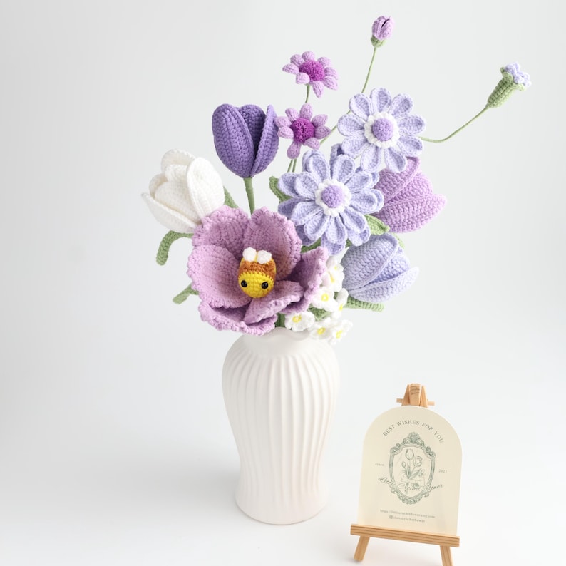 May include: A bouquet of crocheted flowers in shades of white, lavender, and purple, featuring tulips, daisies, and a small yellow and brown bee in the center. The flowers are arranged in a white ribbed vase next to a small wooden easel with a card.
