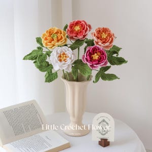 K&ouml;nnte beinhalten: Ein Strau&szlig; geh&auml;kelter Blumen in einer cremefarbenen Vase. Die Blumen sind in Gelb-, Orange-, Rot-, Rosa- und Wei&szlig;t&ouml;nen gehalten, mit gr&uuml;nen Bl&auml;ttern. Der Text "Little Crochet Flower" ist sichtbar. Ein Buch und eine Karte liegen auf dem Tisch.