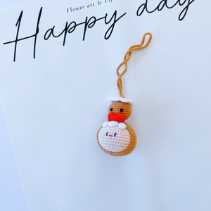 May include: A crocheted gingerbread person ornament with a red bow and white icing. The ornament is hanging from a string and the text "Happy day" is written on a white background.