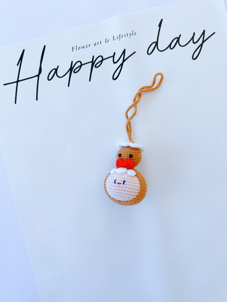 May include: A crocheted gingerbread person ornament with a red bow and white icing. The ornament is hanging from a string and the text "Happy day" is written on a white background.