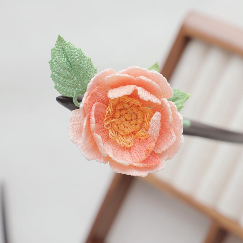 May include: A peach-colored, crocheted flower hair stick with a green leaf. The flower has a detailed, layered design with a darker orange center. The hair accessory is set against a blurred wooden background.