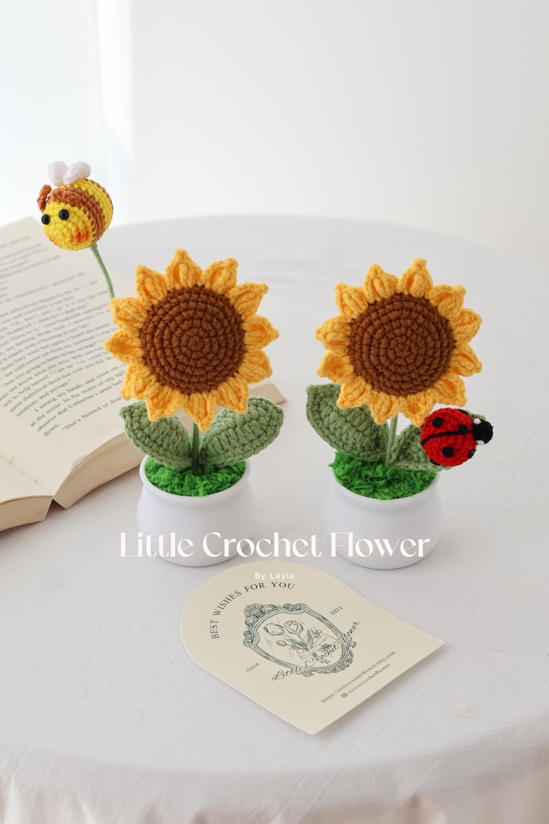 May include: Crocheted sunflower and bee decorations in white pots. The sunflowers have yellow petals and brown centers. A red and black ladybug is attached to one. The text "Little Crochet Flower" is visible.