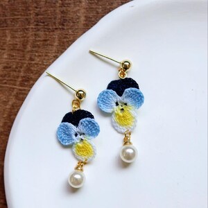 May include: A pair of delicate earrings featuring crocheted blue, yellow, and white flowers with a pearl accent.