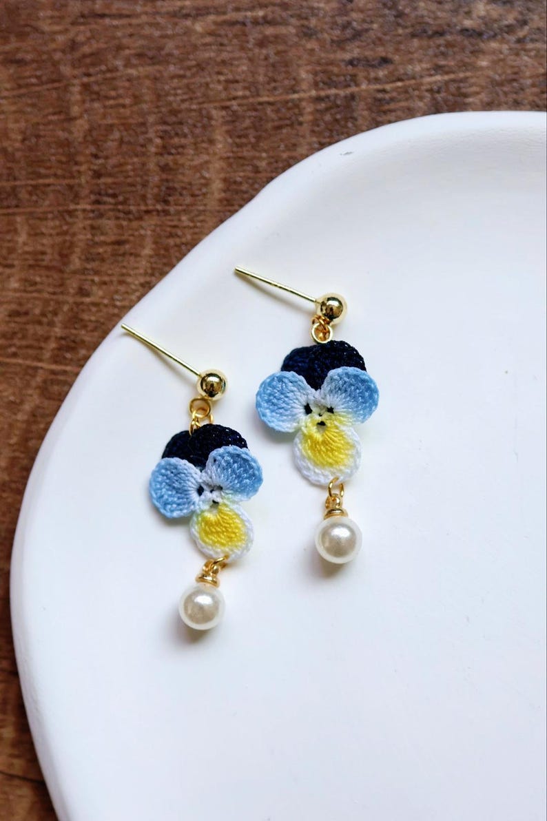 May include: A pair of delicate earrings featuring crocheted blue, yellow, and white flowers with a pearl accent.