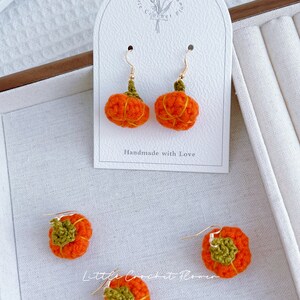 May include: A pair of crocheted pumpkin earrings with gold hooks. The earrings are handmade and feature a green stem and orange pumpkin body. The earrings are displayed on a white card with the text "Handmade with Love" and "Little Crochet Flower".