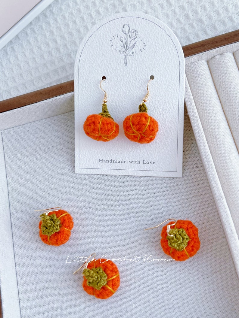 May include: A pair of crocheted pumpkin earrings with gold hooks. The earrings are handmade and feature a green stem and orange pumpkin body. The earrings are displayed on a white card with the text "Handmade with Love" and "Little Crochet Flower".