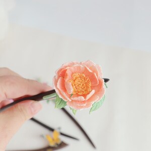 May include: A handcrafted hair stick featuring a peach-colored embroidered flower with a yellow center and green leaves. The flower is attached to a dark wooden stick, creating a unique hair accessory.