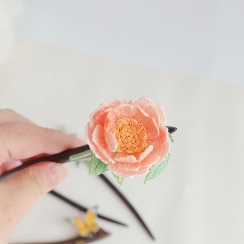 May include: A handcrafted hair stick featuring a peach-colored embroidered flower with a yellow center and green leaves. The flower is attached to a dark wooden stick, creating a unique hair accessory.