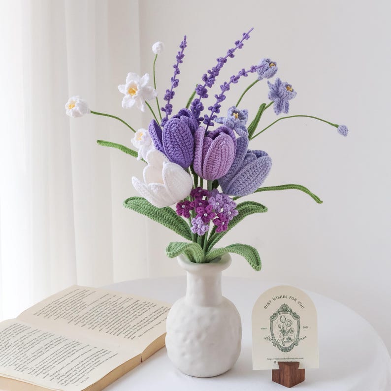 May include: A bouquet of crocheted flowers in shades of white, lavender, and purple, featuring tulips, lavender sprigs, and small blossoms with green stems and leaves, arranged in a textured white ceramic vase next to an open book.
