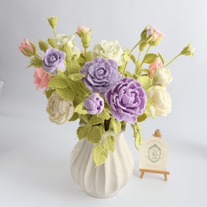 May include: A bouquet of crocheted flowers in shades of lavender, cream, and soft pink, featuring detailed petals and green leaves. The flowers are arranged in a white, ribbed ceramic vase. A small wooden easel displays a card with floral art.