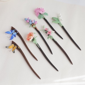 May include: A selection of handcrafted hair sticks, each adorned with floral and butterfly embellishments. The wooden sticks showcase diverse designs, including pink hydrangea, blue berries, and a yellow butterfly. Each stick is unique and detailed.