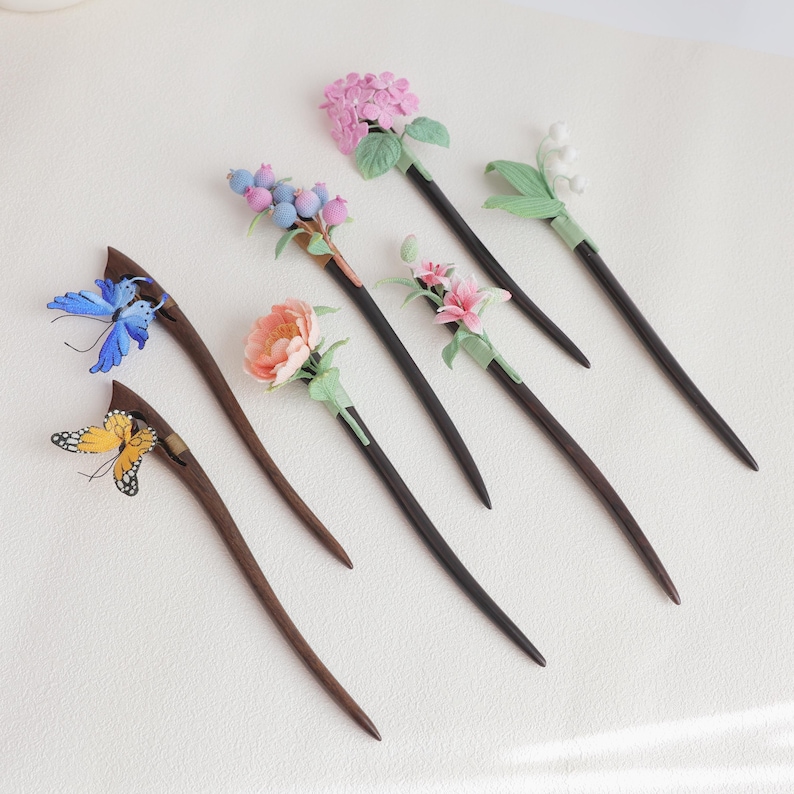 May include: A collection of handcrafted hair sticks with decorative floral and butterfly accents. The wooden sticks feature various designs, including pink hydrangea, blue berries, and a yellow butterfly. Each stick is unique and detailed.