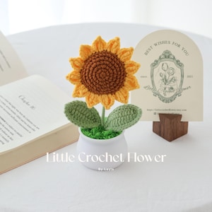 May include: A handmade crochet sunflower in a white pot with green leaves and faux grass. The sunflower has yellow petals and a brown center. A card with the text "Best Wishes For You" is in the background. The text "Little Crochet Flower" is at the bottom.