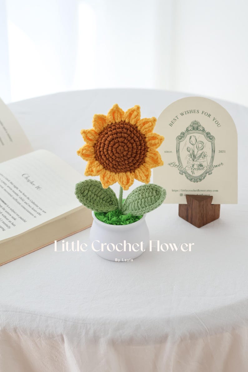 May include: A handmade crochet sunflower in a white pot with green leaves and faux grass. The sunflower has yellow petals and a brown center. A card with the text "Best Wishes For You" is in the background. The text "Little Crochet Flower" is at the bottom.