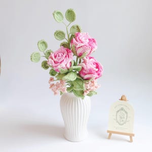 May include: A crochet flower arrangement with three pink roses, green leaves and stems, in a white vase. The arrangement includes small pink flower buds and a small card on a wooden easel.