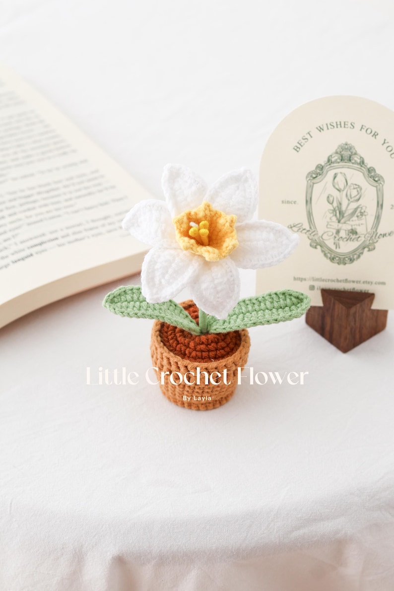 K&ouml;nnte beinhalten: Handgefertigte H&auml;kelblume in einem kleinen Topf. Die wei&szlig;e Blume hat eine gelbe Mitte und gr&uuml;ne Bl&auml;tter. Der Topf ist braun. Der Text "Little Crochet Flower" ist sichtbar.