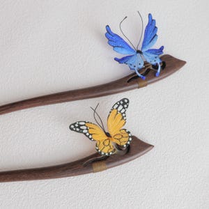 May include: Two wooden hair sticks with butterfly accents. One stick features a blue butterfly, the other an orange butterfly with black and white markings. The hair accessories are set against a white background.