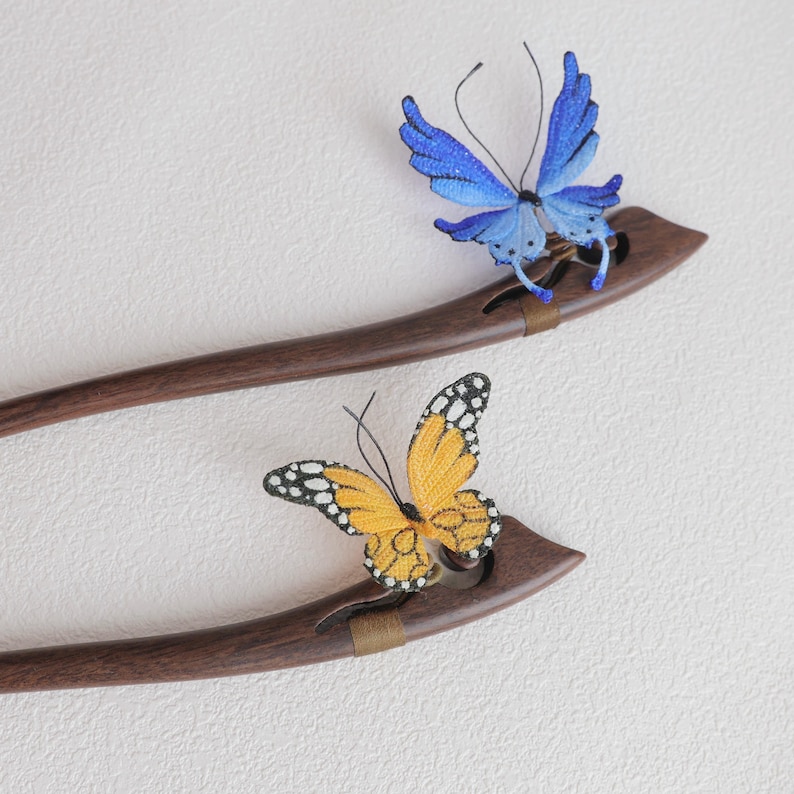 May include: Two wooden hair sticks with butterfly accents. One stick features a blue butterfly, the other an orange butterfly with black and white markings. The hair accessories are set against a white background.