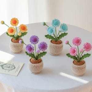 Puede incluir: Cuatro arreglos florales de ganchillo en macetas beige pequeñas. Cada maceta contiene una flor de diferente color: amarillo y naranja, azul, morado y rosa. Las flores tienen hojas verdes y están dispuestas sobre una superficie blanca.