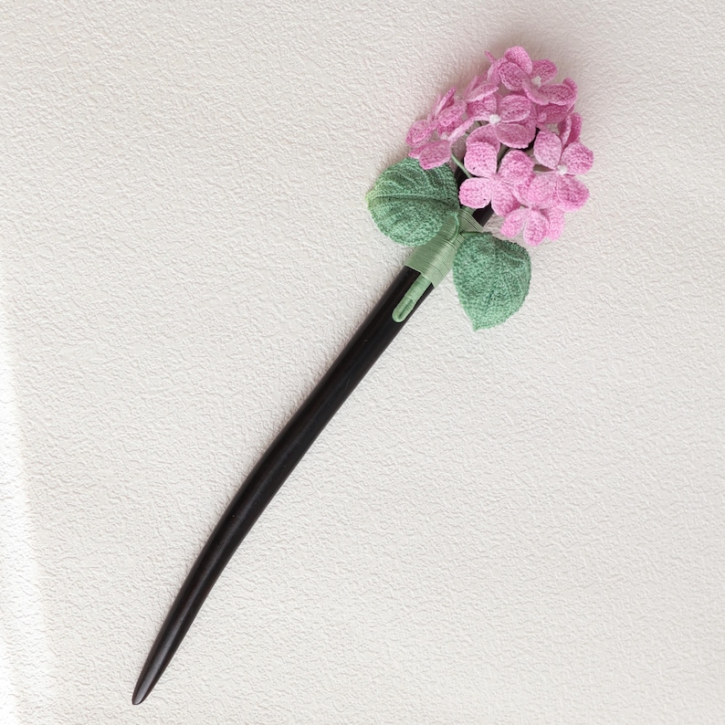 May include: A black wooden hair stick with a decorative pink hydrangea flower and green leaves. The flower is made of a textured fabric, and the leaves are a light green color. The hair accessory is a traditional hair ornament.