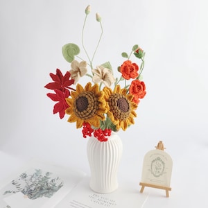 May include: A bouquet of crocheted flowers in a white ribbed vase, featuring two yellow sunflowers with brown centres, red maple-like leaves, orange roses, beige blossoms, and small red berries on green stems, with a small easel sign nearby.