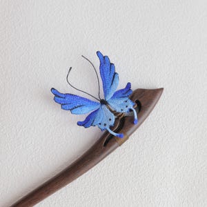 May include: A wooden hair stick with a blue butterfly accent. The butterfly has intricate detailing with shades of blue and black. The hair accessory is a dark brown color.