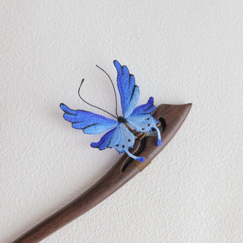 May include: A wooden hair stick with a blue butterfly accent. The butterfly has intricate detailing with shades of blue and black. The hair accessory is a dark brown color.